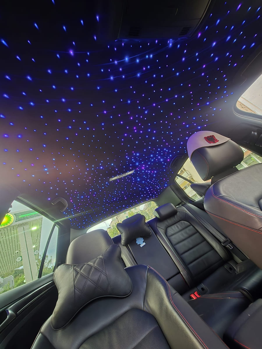 Fertiger Sternenhimmel im Dachhimmel eines Fahrzeugs, aufgenommen bei Tageslicht, durch das Seitenfenster sind eine Tankstelle und Bäume zu sehen.
